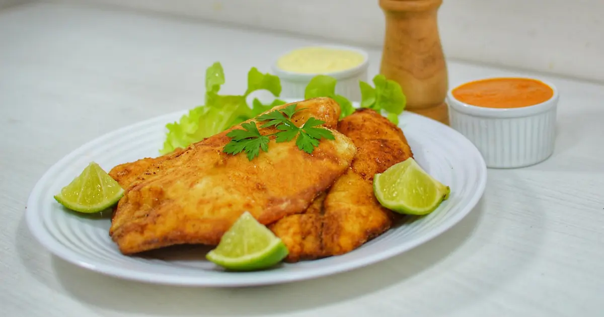 Crispy pan-fried fish fillets served on a white plate with fresh lime wedges, lettuce, and dipping sauces.