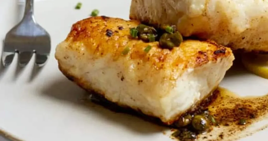 Pan-seared ling cod fillet with crispy golden crust, drizzled with butter and garnished with fresh parsley and lemon slices.